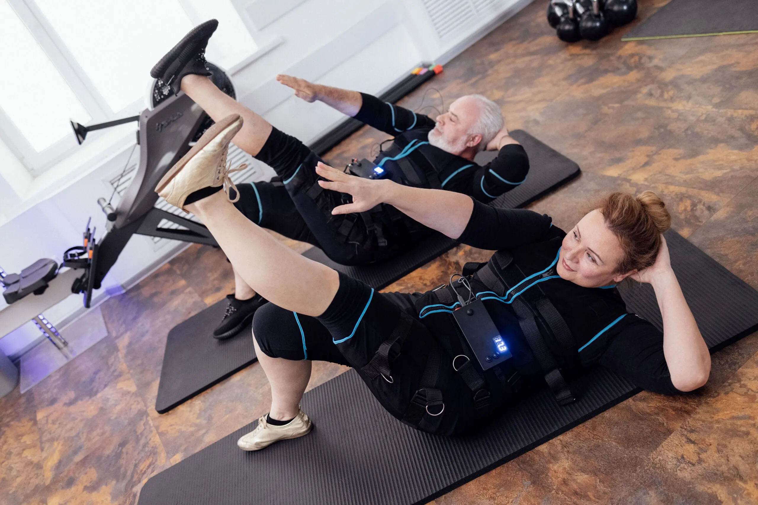 Elderly couple of people in ems suits pump oblique abdominal muscles on floor in gym